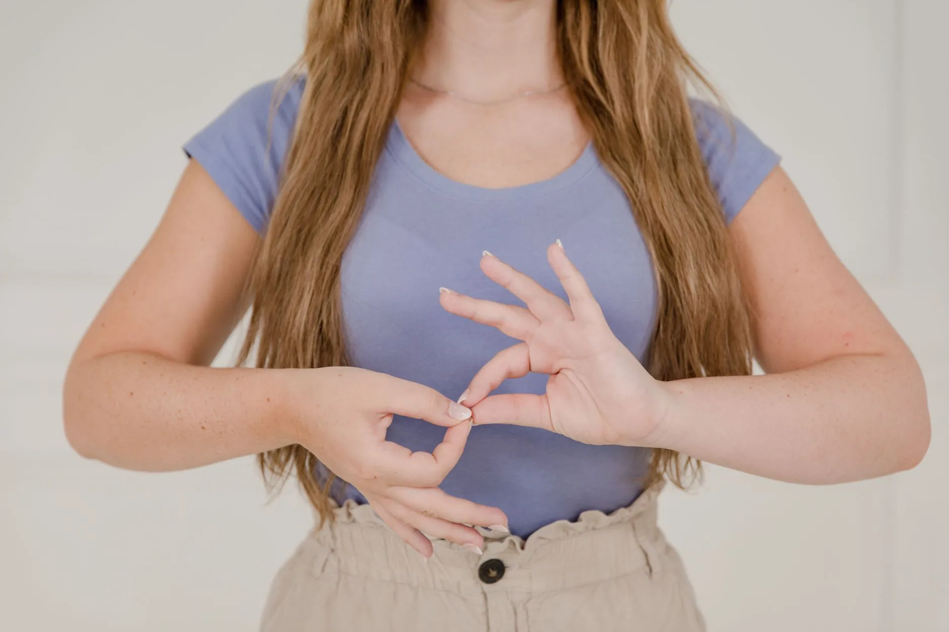 Femme aux longs cheveux en t-shirt bleu faisant un signe de langue des signes devant un mur blanc