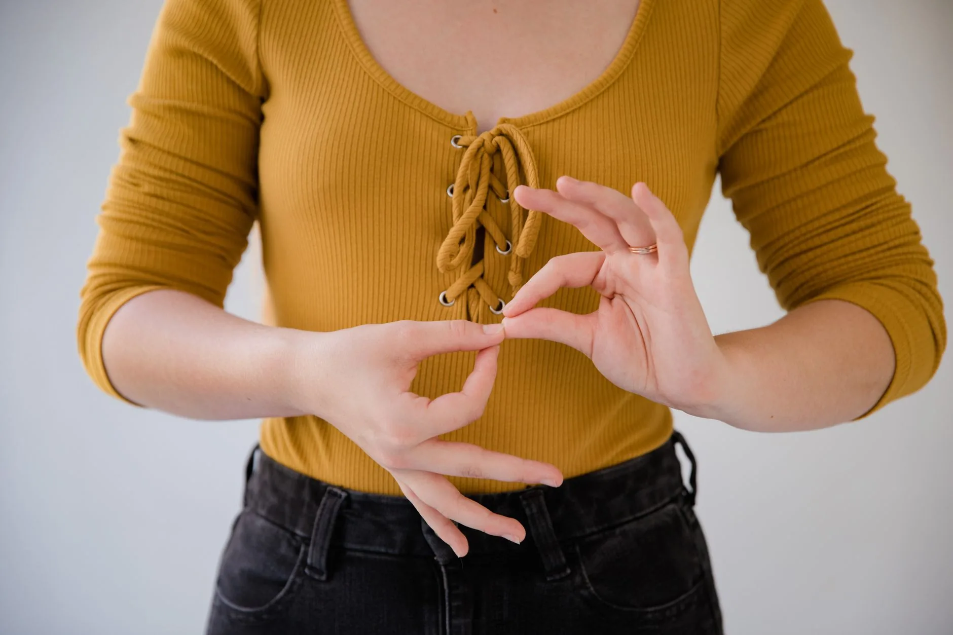 Personne en haut jaune moutarde et jeans noirs faisant un geste des mains sur fond gris