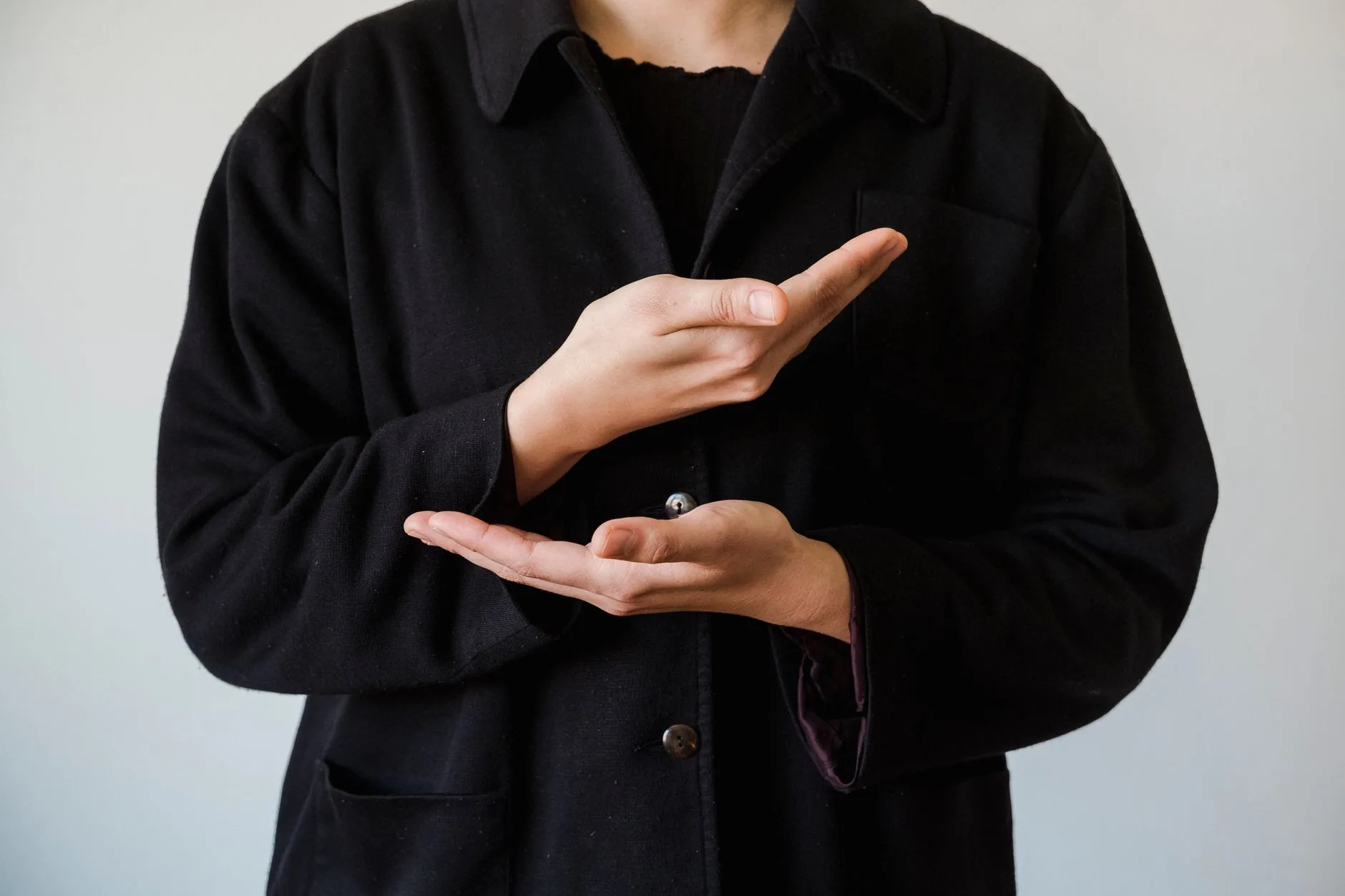 Mains d'une personne en manteau noir formant un geste devant le torse sur fond gris clair